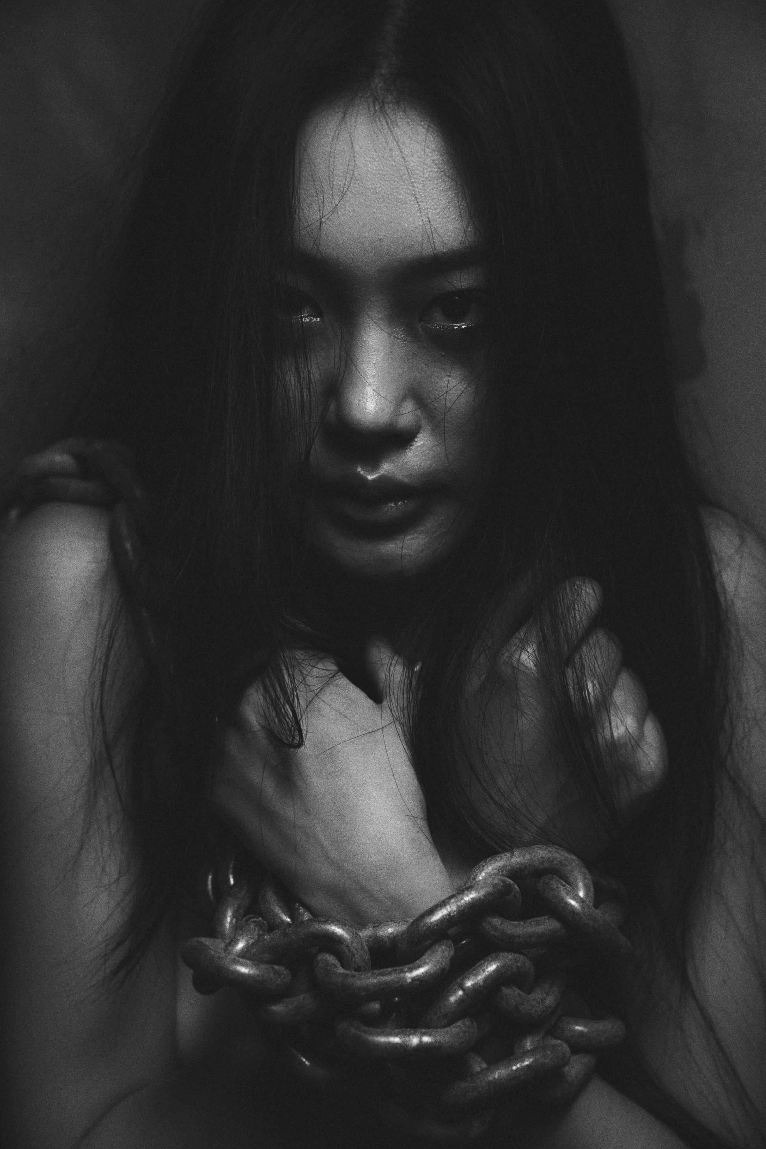 A black-and-white portrait. The subject looks directly into the camera, her arms crossed close to her chest and hands near her shoulders, with a thick chain in front of her wrists.